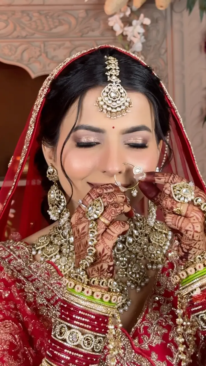 A bride in a red bridal lehenga touching her nose ring, henna-covered hands, layered kundan jewellery and bangles, an ornate carved doorway behind her.